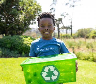 Front view of a smiling young African American boy standing in a field wearing a t shirt with volunteer written on it, holding a green plastic recycling crate and smiling to camera